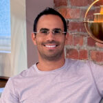 A smiling man wearing glasses and a pink t-shirt, sitting in a lounge in front of exposed brick.