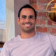 A smiling man wearing glasses and a pink t-shirt, sitting in a lounge in front of exposed brick.