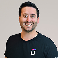 Smiling man with short dark hair and a beard, wearing a black T-shirt with a logo featuring a colorful graphic and the letter U, standing against a neutral background.
