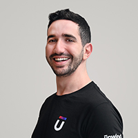 Smiling person with short dark hair and a beard, wearing a black shirt with a logo that includes the letter U. The background is a plain light grey.