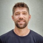 A man with short brown hair and a beard smiles at the camera. He is wearing a dark t-shirt and standing against a neutral, light-colored background.