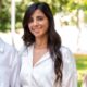 A woman with long, dark hair stands outdoors, wearing a white shirt and subtle jewelry. She is smiling at the camera. The background is blurred, with green foliage and bright lighting.