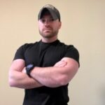 A person wearing a dark t-shirt and a baseball cap stands with arms crossed. The background is a plain, light-colored wall.