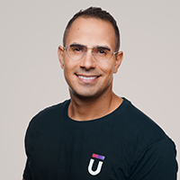 Smiling man with short hair, wearing glasses and a dark T-shirt with a logo on it, stands against a plain light background.