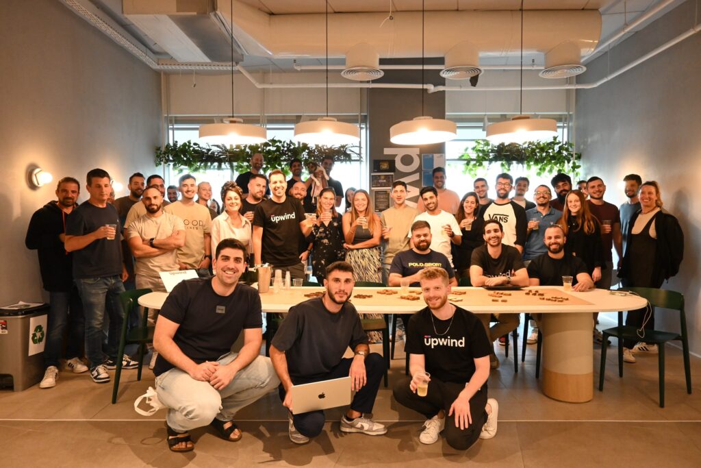 A large group of people pose and smile in a modern office space, with some standing and others sitting at a table. Four people in front wear matching upwind shirts, and plants hang in the background.