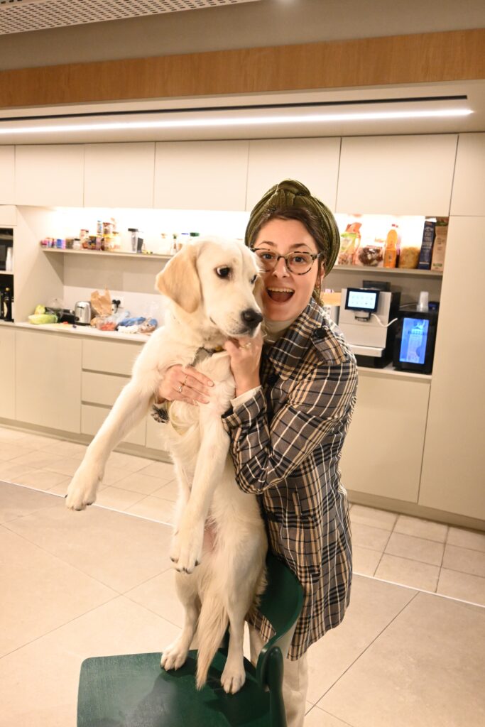 A smiling person in glasses and a headscarf holds a large, light-colored dog standing on a green chair in a modern kitchen with shelves and appliances in the background.