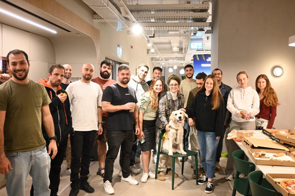 A group of people smiling and posing together indoors, with pizzas on tables nearby. One person is sitting on a chair with a happy dog beside them. The environment appears to be casual and friendly.