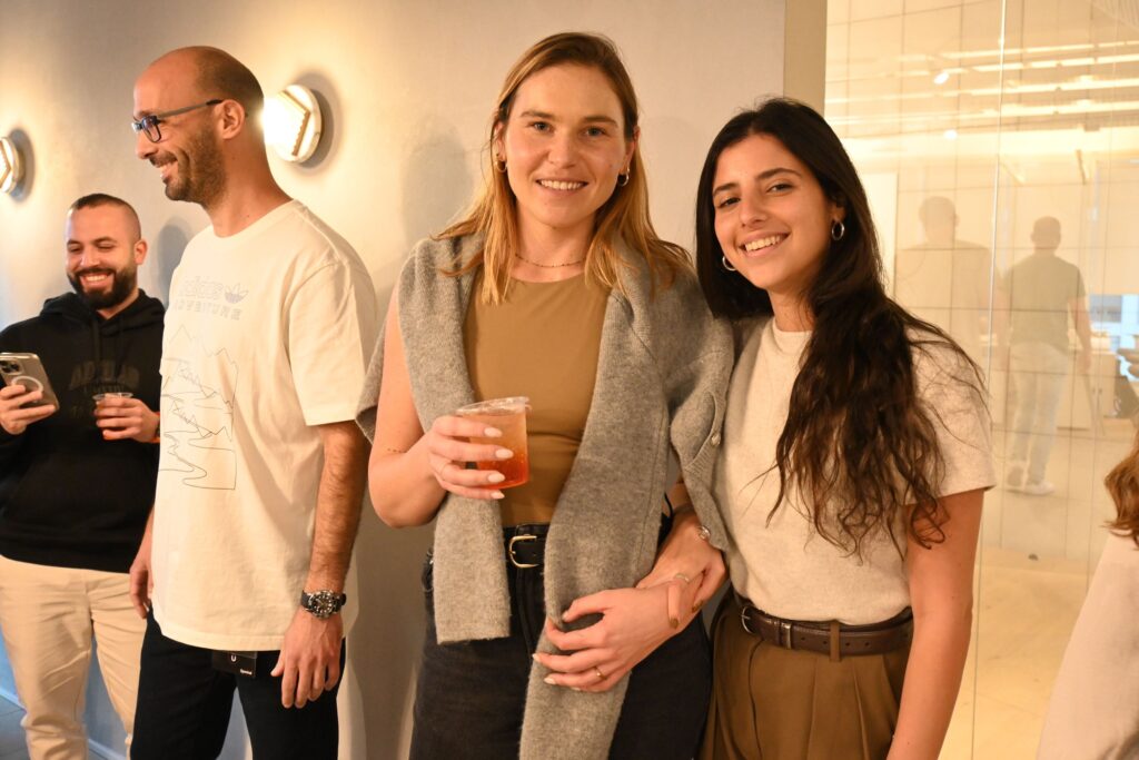 Four people stand together indoors, smiling and chatting. Two women in the center are posing arm-in-arm, one holding a drink. The other two people, both men, are standing nearby, one laughing and the other looking away.