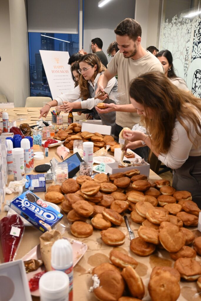 A group of people gathers around a table covered with many pastries, decorating them with whipped cream, icing, and various toppings in a bright, modern room. Supplies and ingredients are scattered across the table.