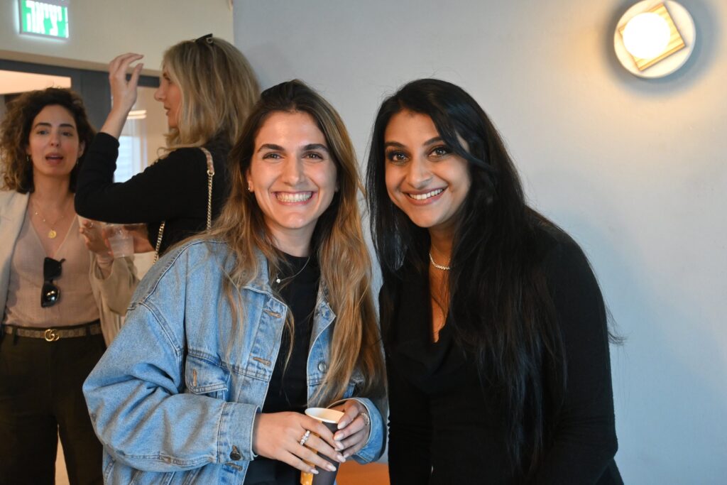 Two women smiling at the camera, one in a denim jacket holding a cup and the other in a black top. In the background, two other women are conversing near a light blue wall with a round light fixture.