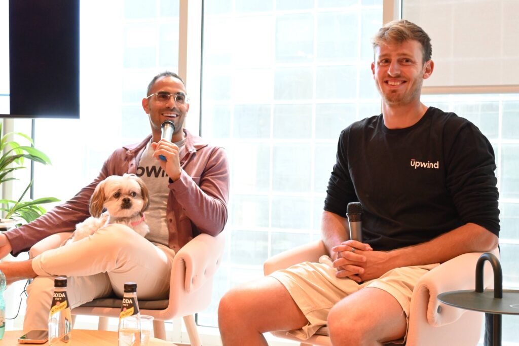 Two men sit on white chairs holding microphones, smiling at an indoor event. One man has a small dog on his lap. Both wear Upwind shirts. There are drinks on the table in front of them, and large windows in the background.