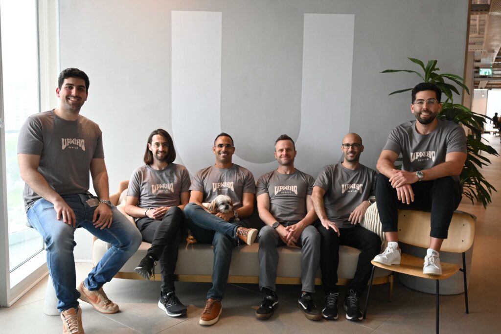 Six men in matching t-shirts sit and smile in a modern office, with a small dog on one person’s lap. They are seated on a couch and bench, with a large plant and natural light coming from the left.