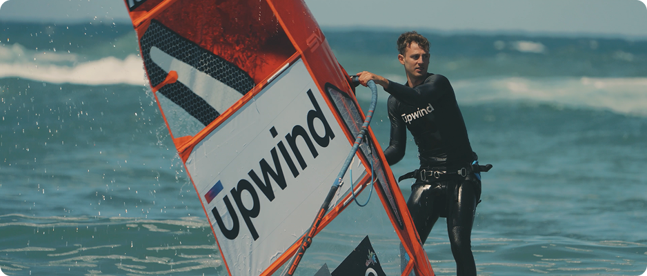 A person in a wetsuit windsurfing on the ocean, holding onto an orange sail labeled Upwind with waves in the background.