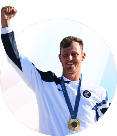 A male athlete wearing a white and navy tracksuit raises his fist in celebration, smiling and displaying a gold medal around his neck.