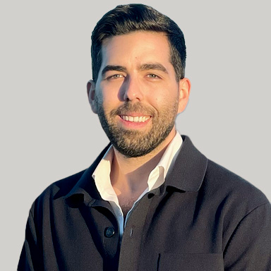 A man with short dark hair and a beard is smiling, wearing a dark jacket over a white shirt, standing against a plain light gray background.
