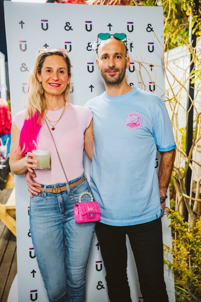 A woman with long blonde and pink hair stands smiling next to a man with a shaved head, both posing in front of a patterned backdrop. She holds a drink and wears a pink purse; he wears a blue shirt and sunglasses on his head.