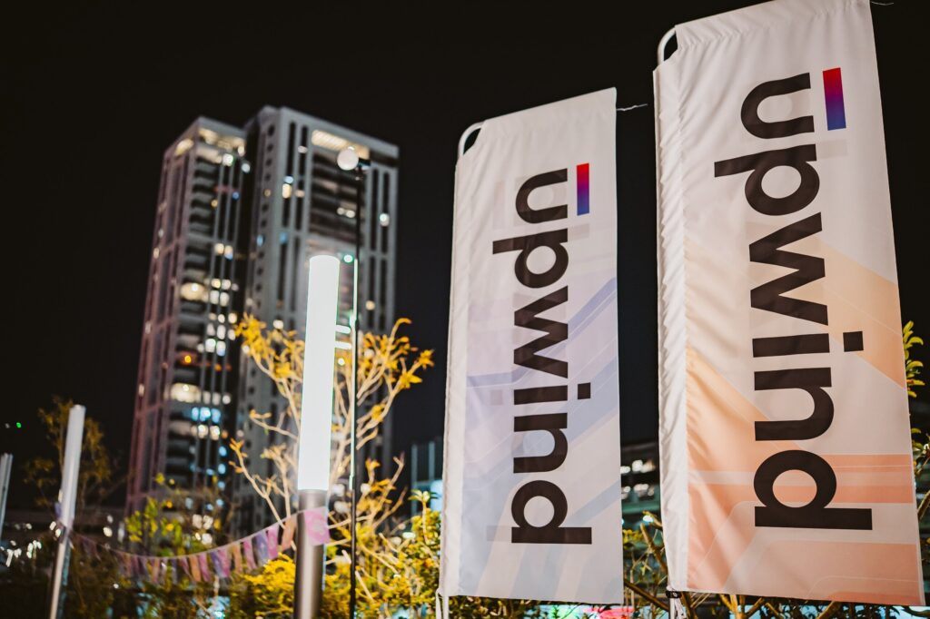 Two tall banners with the text “upwind” stand outdoors at night, with a lit lamp post, some plants, and modern high-rise buildings visible in the background.
