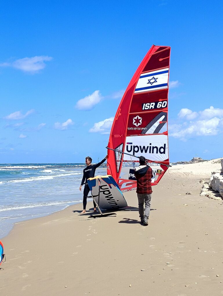 Two people walk along a sandy beach carrying a red and white windsurfing sail with the Israeli flag and the word Upwind on it. The ocean and blue sky with clouds are in the background.