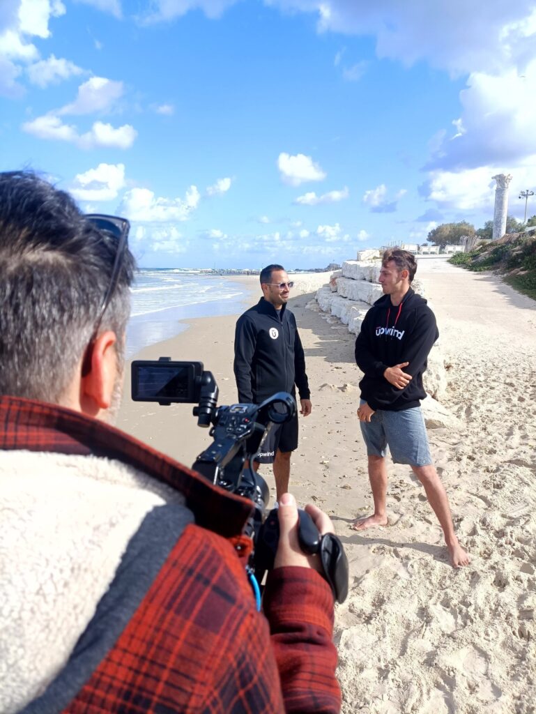 A person with a camera films two men standing and talking on a sandy beach near the water, with rocks and blue sky with clouds in the background.
