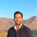 A man with dark hair and a beard, wearing a dark jacket over a white shirt, stands outdoors smiling with mountains and a clear blue sky in the background.